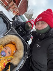 Talvinen ulkokuva, jossa aikuinen ja kaksi lasta ovat ulkona lumisessa pihassa. Aikuinen pitää selfie-kameralla kuvaa ja käyttää punaista pipoa sekä mustaa talvitakkia. Vieressä seisoo lapsi talvivaatteissa ja kasvot osittain peitettynä. Etualalla on vauva lämpimästi puettuna rattaissa, keltainen haalari ja tutti suussa. Taustalla näkyy punainen puutalo ja parkkeerattu auto.