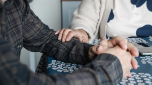 Lähikuva kahden ihmisen käsistä pöydän ääressä. Toinen henkilö pitää toisen kättä omiensa välissä, ja toinen koskettaa lohduttavasti toisen käsivartta.