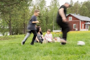 Perhe viettää aikaa ulkona maaseudulla: lapset ja aikuinen pelaavat palloa vihreällä nurmella, ja taustalla näkyy punainen talo sekä kesäinen maisema.