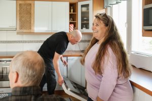 Kolme ihmistä keittiössä: nainen seisoo ja hymyilee, yksi henkilö avaa astianpesukonetta ja kolmas istuu pöydän ääressä.