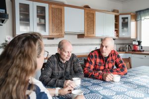 Kuvassa kolme ihmistä istuu keittiön pöydän ääressä. Kaksi miestä ja yksi nainen keskustelevat keskenään kahvikuppien äärellä. Nainen näkyy kuvassa sivusta etualalla, ja miehet istuvat vastapäätä häntä. Taustalla on vaalea keittiö kaapistoineen ja kodinkoneineen, ja tunnelma vaikuttaa rauhalliselta ja keskittyneeltä.